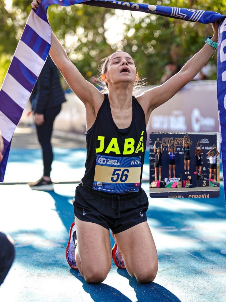 A corredora, com os braços erguidos e segurando uma bandeira listrada de azul e branco, ajoelha-se em êxtase no chão. Seu rosto está voltado para cima, com os olhos fechados e a boca aberta em um grito de alegria ou exaustão. Ela veste um top preto com a palavra "JABÁ" em letras amarelas e um número de peito "56", indicando que ela é uma atleta de elite. Shorts pretos e tênis vermelhos e brancos completam seu traje de corrida. Ao fundo, em um pequeno quadrado sobreposto, mostra-se um pódio com quatro mulheres celebrando suas vitórias, com sacolas de prêmios e troféus. A imagem transmite a emoção e a conquista de uma vitória em uma corrida.