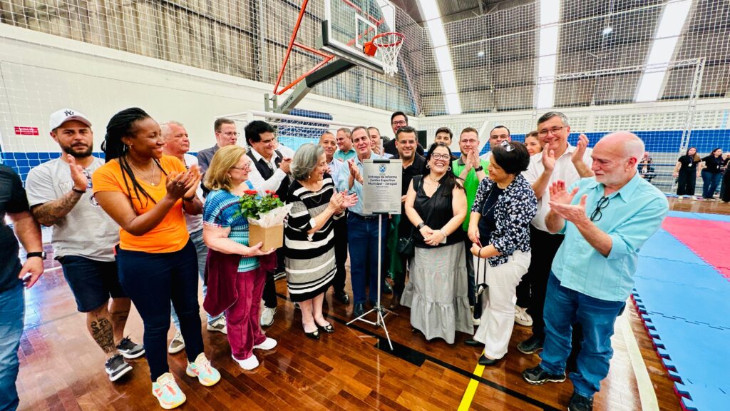 A imagem mostra um grupo de 10 pessoas, 8 homens e 2 mulheres, posando para uma foto em um ambiente interno que parece ser uma quadra esportiva, com uma rede de gol ao fundo.A mulher no centro, segurando um pequeno arranjo de flores vermelhas, está vestida com um vestido listrado em preto e branco. Ela está cercada por vários homens em diferentes trajes casuais e semi-formais. A iluminação sugere que a foto foi tirada em um ambiente bem iluminado, possivelmente com luzes de teto. A maioria das pessoas está sorrindo e olhando para a câmera, indicando um momento de celebração ou reunião.