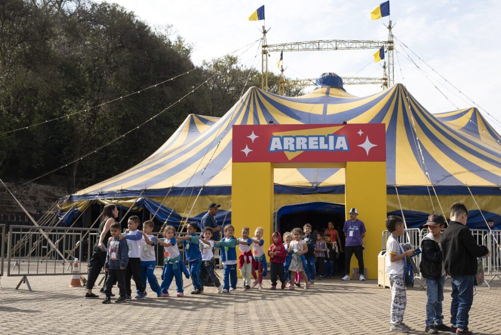 Crianças saem em fila indiana da lona de circo Arrelia acompanhadas por monitores.