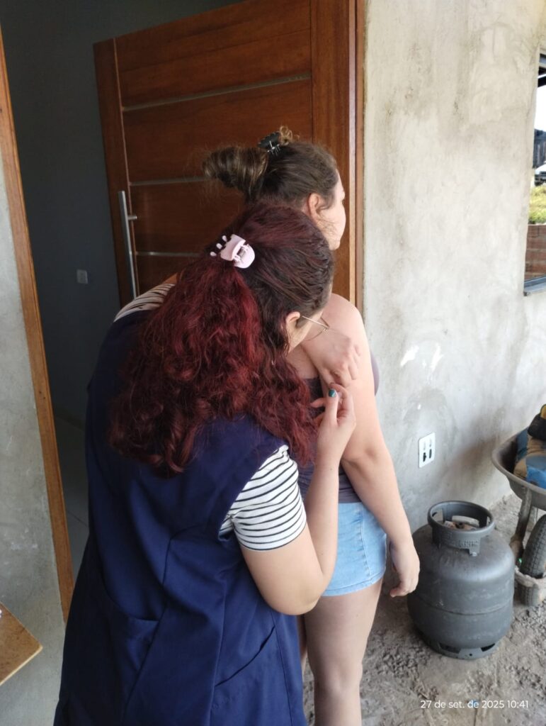 A imagem mostra duas mulheres em pé em frente a uma porta de madeira. A mulher da frente, com cabelos longos e ruivos presos em um rabo de cavalo com um prendedor rosa, está usando um colete azul escuro sobre uma blusa listrada de branco e preto. Ela está de costas para a câmera e parece estar aplicando uma injeção no braço da outra mulher. A mulher de trás, com cabelos castanhos presos em um coque com um prendedor preto, está de perfil, olhando para a direita. Ela está usando uma blusa cinza e shorts jeans. O fundo da imagem mostra uma parede de concreto grosseiro, uma janela e um botijão de gás cinza. Há também uma carretilha com alguns objetos em cima, parcialmente visível à direita. No canto inferior direito da imagem, há uma marcação de data e hora: "27 de set. de 2025 10:41".