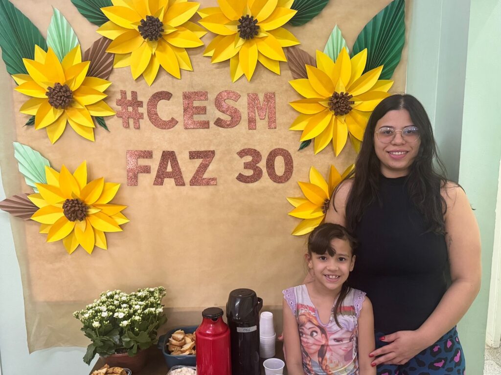 A imagem mostra duas pessoas, uma mulher adulta e uma menina, em frente a um banner decorado com girassóis de papel e as palavras "#CESM FAZ 30" em letras brilhantes. A mulher, à direita, usa um top preto e óculos redondos, sorrindo para a câmera. A menina, à esquerda, veste uma camiseta com estampa de personagens e também sorri. O banner tem um fundo marrom claro, com grandes girassóis amarelos feitos de papel, intercalados com folhas verdes e marrons. As palavras "#CESM FAZ 30" estão escritas em glitter rosa. Na parte inferior da imagem, uma mesa está parcialmente visível, com uma planta em vaso com pequenas flores brancas, uma garrafa térmica vermelha, uma garrafa térmica preta com a inscrição "Chá" e copos plásticos brancos. Há também recipientes com alimentos, como pães ou torradas. O ambiente parece ser de uma celebração ou evento.