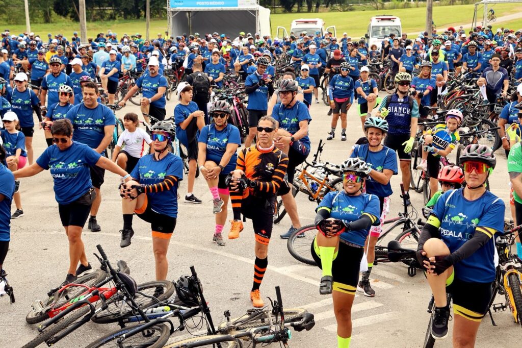 A imagem mostra um grande grupo de ciclistas reunidos ao ar livre, possivelmente antes de um evento ou passeio de bicicleta. A maioria das pessoas está vestindo camisetas azuis com o logotipo "PEDALA TOUR" e algumas usam capacetes de ciclismo. Eles parecem estar se alongando, com muitos indivíduos realizando exercícios de aquecimento, como levantar uma perna em direção ao peito ou esticar os braços. Ao fundo, há uma tenda com a inscrição "POSTO MÉDICO", indicando que se trata de um evento organizado. Várias bicicletas estão estacionadas ou apoiadas no chão, e algumas vans ou veículos de apoio também são visíveis. O ambiente é ensolarado e parece ser um dia agradável para atividade ao ar livre. A multidão é diversa, com homens, mulheres e até algumas crianças presentes.