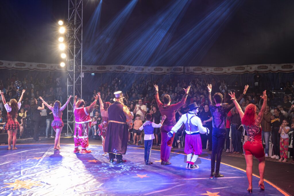 Vários artistas de circo de costas levantam os braços no palco para público lotado.