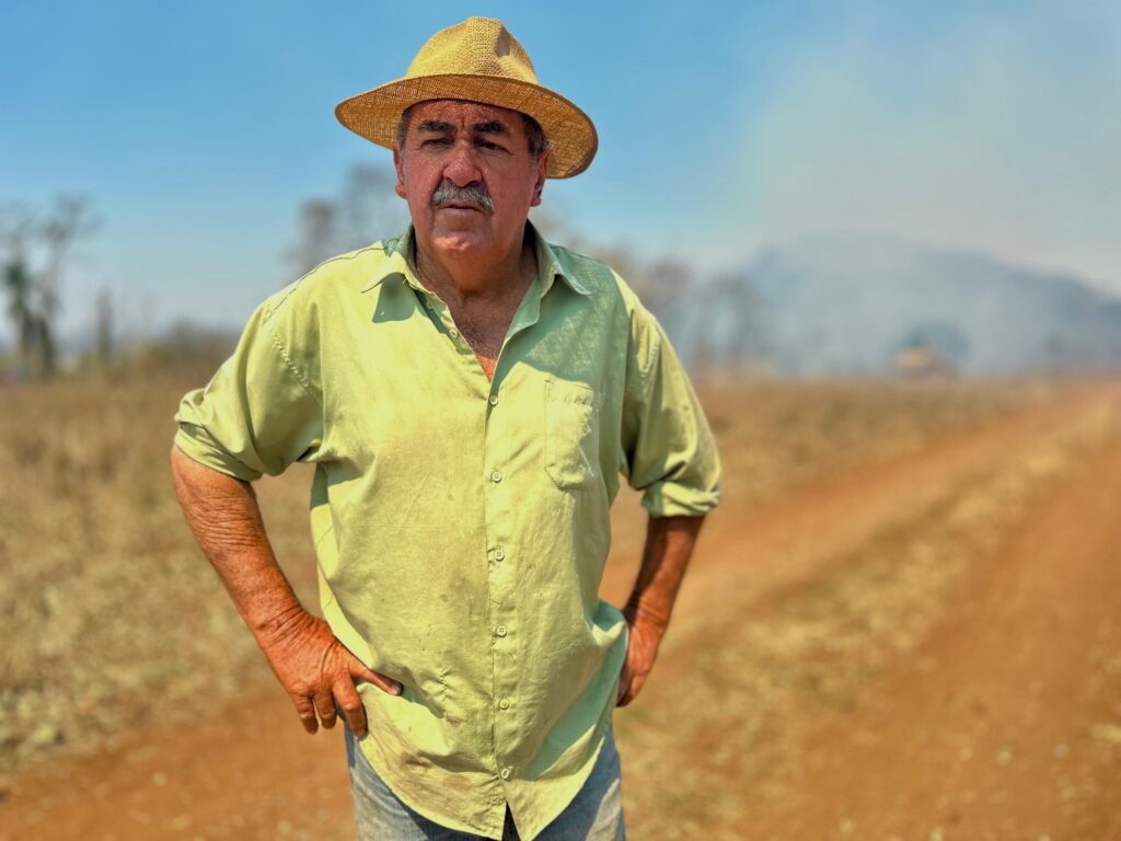 Um fazendeiro de meia-idade, com um chapéu de palha e uma camisa de botões verde-claro, está em pé em um campo seco com uma estrada de terra ao fundo. Ele tem as mãos nos quadris e olha diretamente para a câmera com uma expressão séria. A fumaça pode ser vista ao longe, sugerindo uma queimada ou alguma outra atividade agrícola na área. O sol brilha intensamente, criando sombras fortes no rosto do homem e destacando as rugas em seus braços.