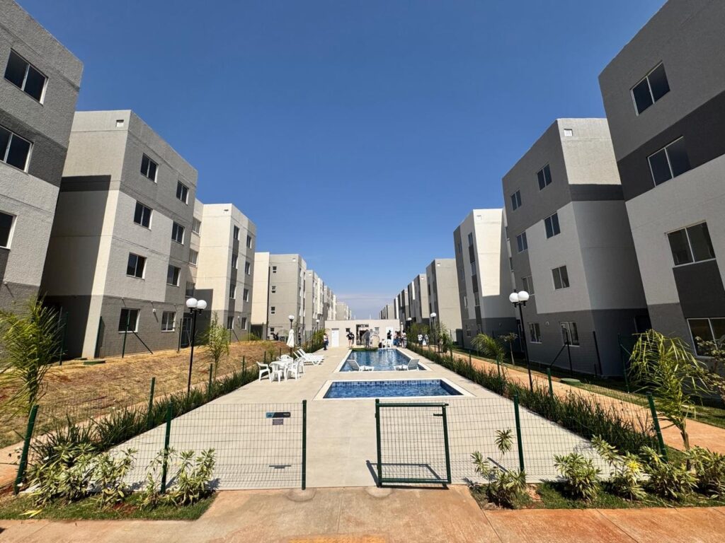 A imagem mostra um complexo de apartamentos modernos, com uma piscina retangular no centro. Os prédios são altos, com vários andares e janelas uniformes, pintados em tons de cinza claro e escuro. Há uma área gramada ao redor da piscina, com algumas cadeiras brancas e guarda-sóis. Uma cerca de metal com um portão se estende pela frente da imagem, separando a área da piscina do caminho de cimento. O céu azul e limpo sugere um dia ensolarado.