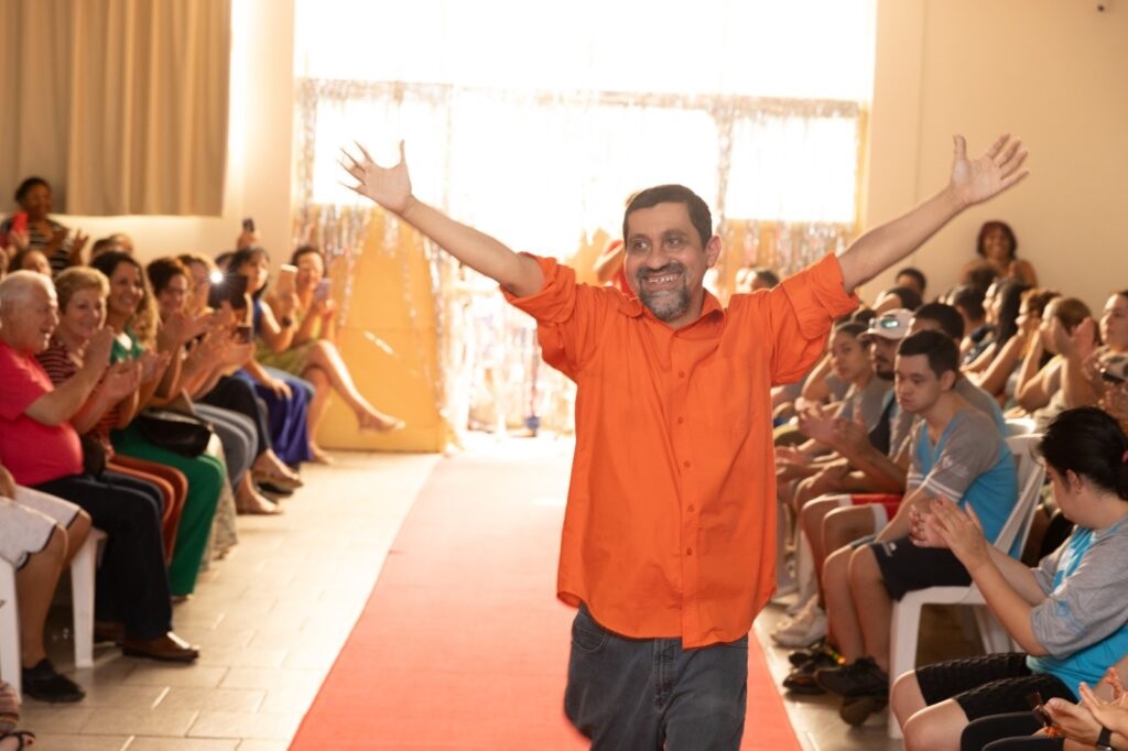 A imagem mostra um homem sorridente no centro, com os braços erguidos em um gesto de celebração ou saudação. Ele veste uma camisa laranja vibrante e calças jeans. Ele está em uma passarela vermelha, que se estende em direção a um público sentado em ambos os lados. O público parece estar aplaudindo e engajado no evento, com muitas pessoas segurando celulares para tirar fotos ou gravar vídeos. O ambiente sugere um evento comunitário ou uma apresentação, possivelmente um desfile de moda ou uma celebração. As pessoas no público são diversas em idade e etnia.