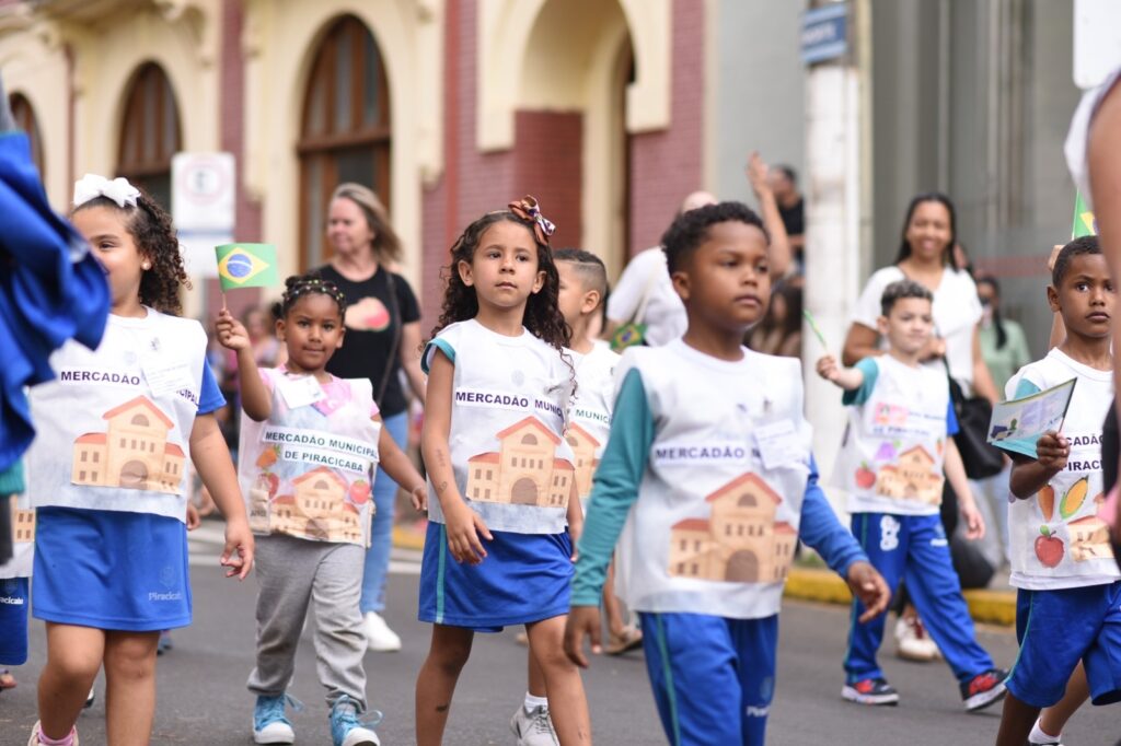 Na imagem aparecem 7 crianças, sendo quatro meninos e três meninas, desfilando em evento cívico por uma rua. Na camiseta que vestem está escrito Mercadão Municipal. O uniforme é branco e azul.