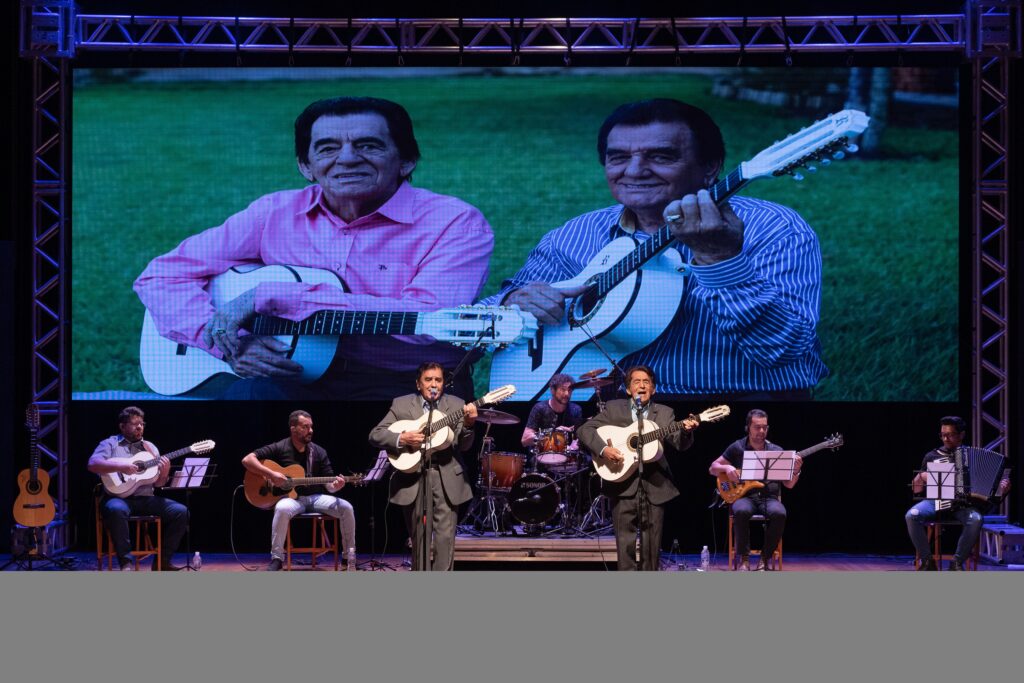 A imagem mostra uma banda se apresentando em um palco. No fundo, uma grande tela exibe uma imagem de dois homens mais velhos tocando violões. Na frente do palco, há vários músicos.No centro, dois homens em ternos cinza estão cantando e tocando violões brancos. À esquerda deles, um homem está sentado tocando um violão. À direita, um homem está tocando um acordeão e outro está tocando um baixo elétrico. Há também um baterista atrás dos músicos da frente.A iluminação é focada nos músicos, com a tela de fundo iluminando o palco. A atmosfera parece ser de um concerto musical.