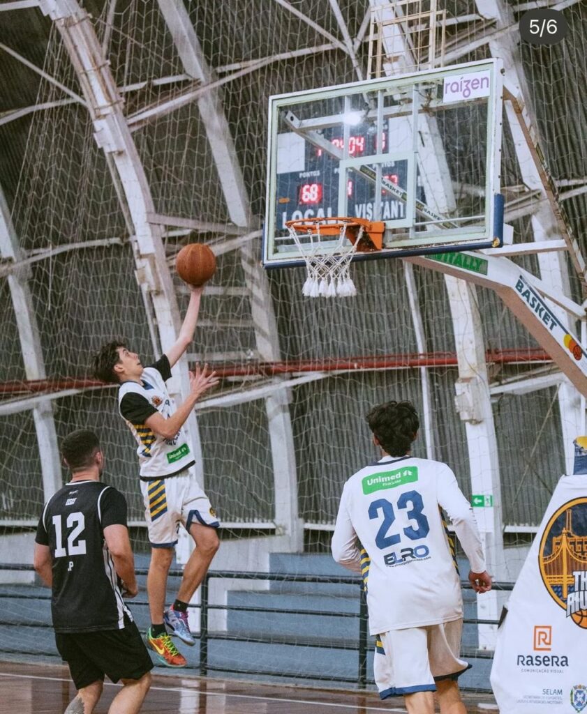 A imagem mostra uma partida de basquete em uma quadra interna. Um jogador com uma camisa branca com listras azuis e amarelas está no ar, prestes a arremessar a bola na cesta. Ele está com a mão direita levantada, segurando a bola, e o olhar focado no aro. Dois outros jogadores estão visíveis na quadra. Um deles, vestindo uma camisa preta com o número 12 em branco, está de costas para a câmera, observando o jogador que arremessa. O outro jogador, com uma camisa branca de mangas compridas e o número 23 em azul, também está de costas, com o nome "ELRO" abaixo do número.
