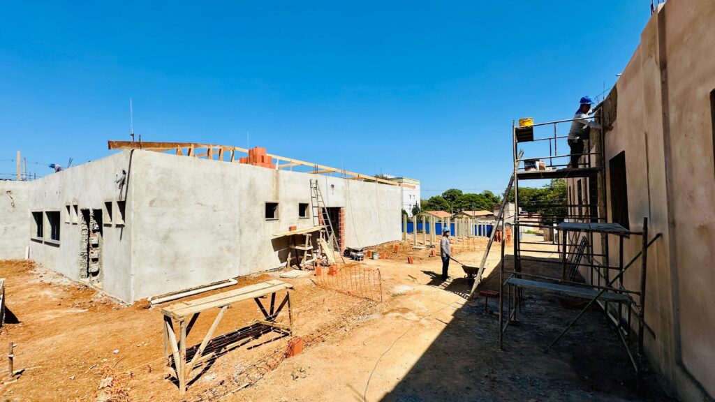 A imagem mostra um canteiro de obras de um edifício em construção, sob um céu azul claro e ensolarado. A estrutura principal do edifício, com paredes de concreto aparente, está em fase de acabamento, com janelas já abertas e alguns detalhes em tijolo aparente. No lado esquerdo da imagem, vemos uma parede com várias janelas e uma abertura que parece ser uma porta ou uma grande janela, com tijolos expostos. Há andaimes de madeira montados em frente a essa parede, com materiais de construção empilhados. Uma escada de metal está apoiada na parede, indicando trabalho em andamento. No centro da imagem, um trabalhador com um capacete azul está em cima de um andaime metálico, aparentemente realizando algum tipo de trabalho de acabamento na parede externa do edifício. Ao lado dele, um carrinho de mão está posicionado no chão de terra batida.