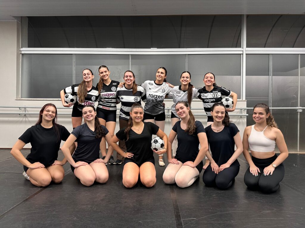 A imagem mostra um grupo de dez jovens mulheres, vestindo uniformes esportivos, em uma sala com piso de madeira escura e uma grande janela ao fundo. As mulheres estão dispostas em duas fileiras, com cinco em cada uma. Na fileira de trás, todas usam uniformes de futebol em preto e branco, com exceção de uma mulher no centro que usa um uniforme branco com detalhes em preto e cinza. Algumas delas seguram bolas de futebol. Seus rostos transmitem alegria e descontração, com sorrisos e poses confiantes. Na fileira da frente, quatro mulheres estão ajoelhadas, vestindo roupas esportivas pretas. Uma delas segura uma bola de futebol. A mulher mais à direita, na fileira da frente, veste um top esportivo branco e calças pretas, e também sorri para a câmera. O ambiente sugere um local de treino ou competição esportiva, possivelmente um ginásio ou estúdio de dança, devido ao piso e às barras de apoio visíveis ao fundo. A iluminação é clara, destacando as figuras das mulheres e suas vestimentas.
