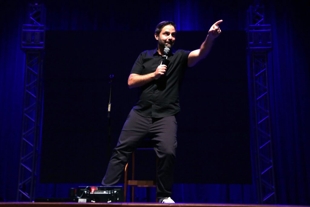 Comediante em pé no palco com microfone aponta para a plateia durante stand-up.
