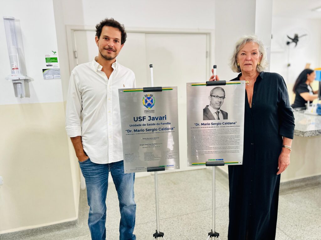 A imagem mostra duas placas comemorativas em um ambiente interno, possivelmente uma unidade de saúde. Duas pessoas, um homem e uma mulher, estão em pé ao lado das placas. O homem, à esquerda, veste uma camisa branca e jeans. Ele tem cabelo castanho encaracolado e barba. Ele está sorrindo levemente para a câmera. A mulher, à direita, veste um vestido preto longo. Ela tem cabelo grisalho curto e usa brincos dourados. Ela está olhando para a câmera com uma expressão neutra. As placas são prateadas e têm texto e imagens. A placa da esquerda tem o brasão de Piracicaba e o texto "USF Javari Unidade de Saúde da Família "Dr. Mario Sergio Caldana"". A placa da direita tem uma foto em preto e branco de um homem de óculos e o mesmo texto. O fundo da imagem mostra uma parede branca, uma porta fechada e parte de um dispensador de lenços de papel ou luvas na parede. Há também uma pessoa desfocada ao fundo, trabalhando em uma mesa.