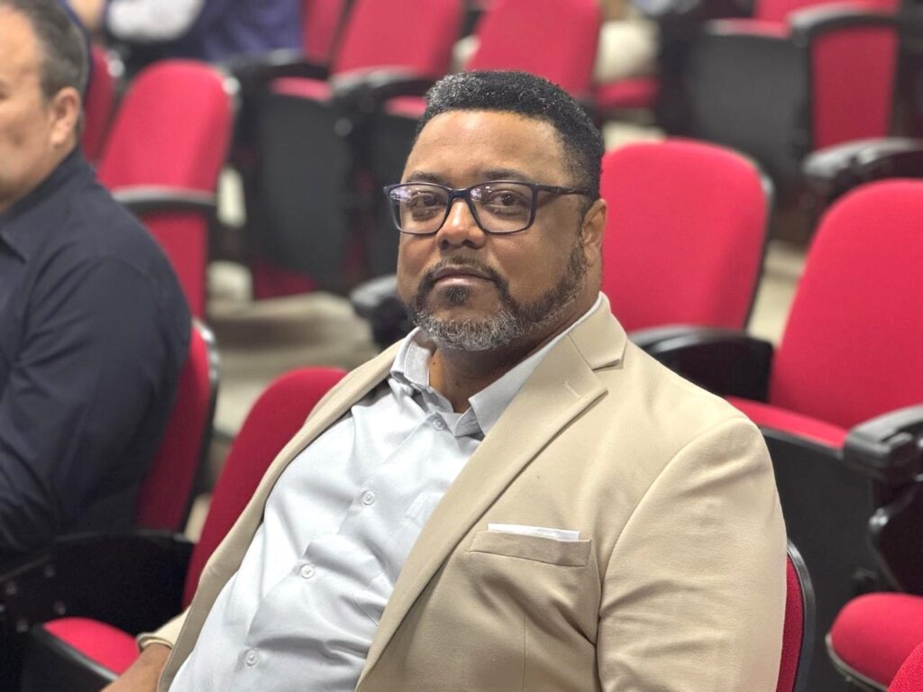 A imagem mostra um homem de meia-idade, com pele morena, usando óculos de aro escuro e um blazer bege sobre uma camisa clara. Ele tem barba e cabelo curto e grisalho. O homem está sentado em uma cadeira em uma sala com várias outras cadeiras vermelhas ao fundo, sugerindo que ele pode estar em um evento, conferência ou auditório. A iluminação é suave, focando principalmente no rosto do homem, que olha diretamente para a câmera com uma expressão neutra.