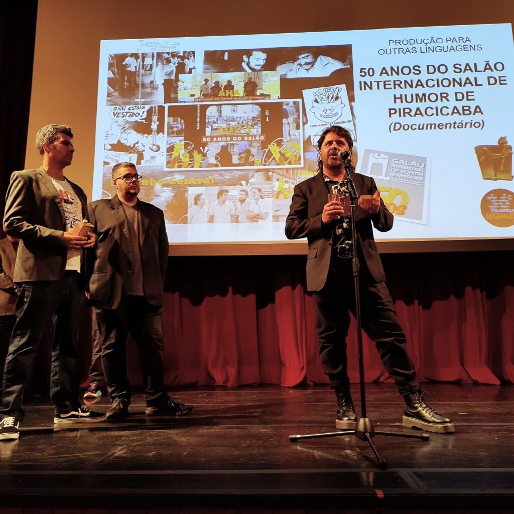 A imagem mostra três homens em um palco, com uma tela ao fundo exibindo uma colagem de imagens e texto. O homem mais à direita está falando ao microfone e segurando um prêmio. Ele está vestindo um terno preto sobre uma camisa estampada e calças pretas, com botas de combate grossas. Os outros dois homens estão em pé ao lado dele, com o homem do meio usando um terno escuro sobre uma camiseta cinza e óculos, e o homem mais à esquerda usando um blazer cinza sobre uma camiseta branca e calças jeans. A tela ao fundo tem o título "50 ANOS DO SALÃO INTERNACIONAL DE HUMOR DE PIRACICABA (Documentário)" e outras imagens relacionadas ao evento. A iluminação do palco é suave, e há cortinas vermelhas atrás da tela.