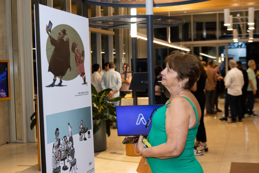 A imagem mostra uma mulher com cabelos castanhos curtos, vestindo um vestido verde vibrante, olhando atentamente para uma exposição de arte. Ela está em primeiro plano, com o corpo ligeiramente virado para a esquerda. Um colar dourado com um pingente delicado adorna seu pescoço. Em sua mão direita, ela segura um celular e um chaveiro amarelo. Ao fundo, outras pessoas estão reunidas, algumas conversando e outras observando as obras expostas. A iluminação ambiente sugere um evento noturno ou em um espaço interno bem iluminado. No centro da imagem, um painel branco exibe duas ilustrações em estilo de desenho animado. A ilustração superior apresenta duas figuras em um círculo, e a inferior mostra um grupo de pessoas sentadas em torno de uma mesa. Um monitor de tela azul com um logotipo branco em forma de "A" é visível ao lado do painel de arte.