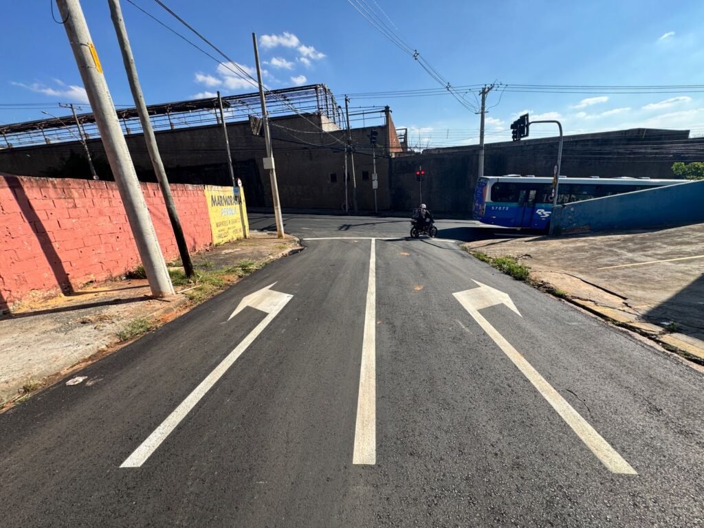 A imagem mostra uma rua com recapeamento recente, evidenciado pelas setas brancas recém-pintadas na via. Há três setas: duas apontando para a esquerda e uma para a direita, indicando as direções permitidas.Ao fundo, um ônibus azul com o número 57073 e o logo "RS" está parado em um cruzamento com semáforo vermelho. Um motociclista com capacete branco e jaqueta preta está se aproximando do cruzamento.À esquerda da rua, uma parede de tijolos vermelhos com um muro baixo e uma placa amarela com a inscrição "MARMORARIA PEROLA" se destacam. Vários postes de luz e fios de eletricidade cruzam o céu azul com algumas nuvens brancas. Ao fundo, uma estrutura metálica e um edifício de concreto completam a paisagem urbana.