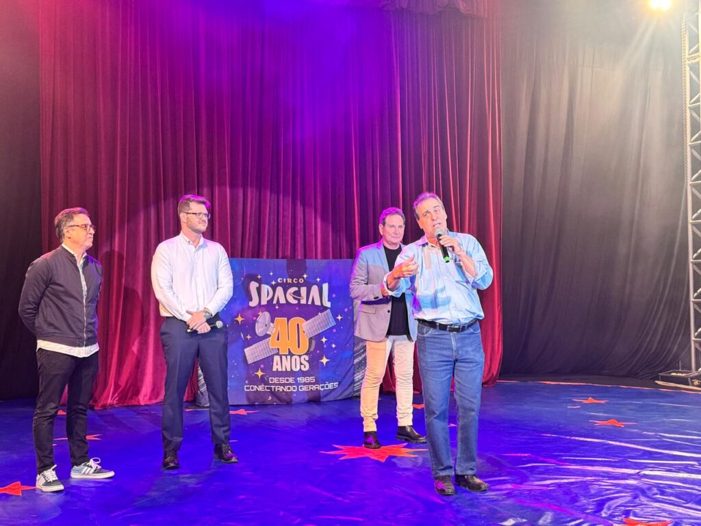 Prefeito fala ao microfone no palco do circo, acompanhado por três autoridades.