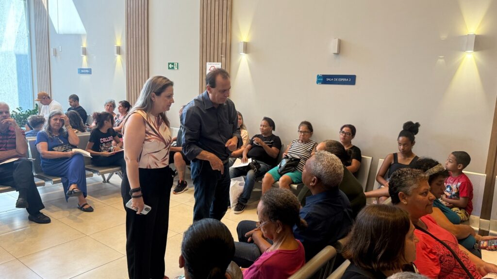 A imagem mostra uma sala de espera com várias pessoas sentadas em cadeiras. No centro, um homem e uma mulher estão conversando. O homem está gesticulando com as mãos enquanto fala, e a mulher está olhando para ele com atenção. Ao fundo, há uma placa com a inscrição "SALA DE ESPERA 2". As paredes são claras e há iluminação embutida no teto. As pessoas na sala de espera parecem estar aguardando atendimento. A maioria está vestida casualmente.