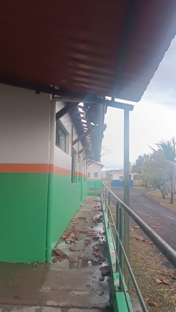 A imagem mostra a lateral de um edifício com a fachada pintada em branco na parte superior e verde na parte inferior, com uma faixa laranja no meio. O telhado de metal cor de ferrugem, que se estende sobre a lateral do edifício, parece danificado, com algumas partes soltas ou quebradas. Restos de telhas de barro estão espalhados no chão, juntamente com poças de água, o que sugere que choveu recentemente ou que houve algum vazamento. Uma grade de metal acompanha a lateral do edifício, protegendo a área externa. Ao fundo, podem ser vistas outras construções e vegetação, incluindo palmeiras, sob um céu nublado.