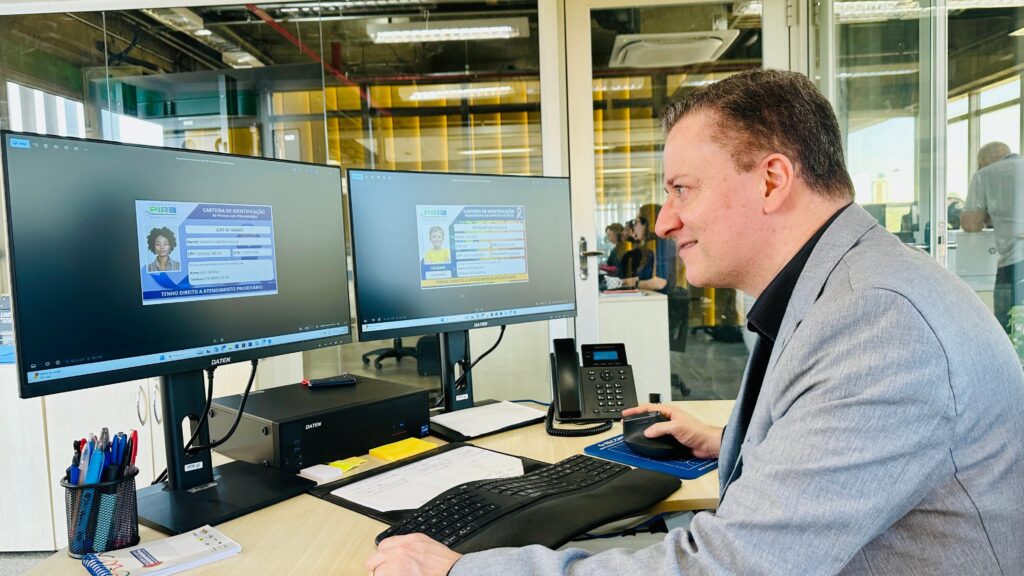 Um homem em um terno cinza está sentado em uma mesa, olhando para dois monitores de computador. Os monitores exibem cartões de identificação. O cartão à esquerda mostra uma mulher com o nome "Valentina da Silva Sousa" e a inscrição "TENHO DIREITO A ATENDIMENTO PRIORITÁRIO". O cartão à direita mostra um menino com o nome "João Souza" e a mesma inscrição. O homem está segurando um mouse e parece estar trabalhando. Há materiais de escritório na mesa, incluindo canetas, um teclado e um telefone. Ao fundo, há paredes de vidro com reflexos de pessoas e do ambiente de escritório.