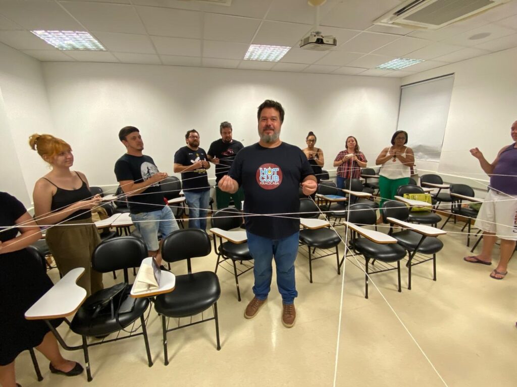 A imagem mostra um grupo de pessoas em uma sala de aula, possivelmente participando de uma atividade de formação ou dinâmica de grupo. Elas estão em pé, conectadas por fios ou cordas que formam uma teia. No centro da imagem, um homem com barba e camisa preta com o logo "HOT CLUB PIRACICABA" está de frente para a câmera, segurando os fios. Ao redor dele, outras pessoas estão distribuídas pela sala, cada uma segurando um fio. As pessoas parecem estar engajadas na atividade, com expressões faciais variadas. A sala de aula está equipada com carteiras e cadeiras, e a iluminação é artificial, vinda de luminárias no teto. O ambiente sugere um local de aprendizado ou treinamento.