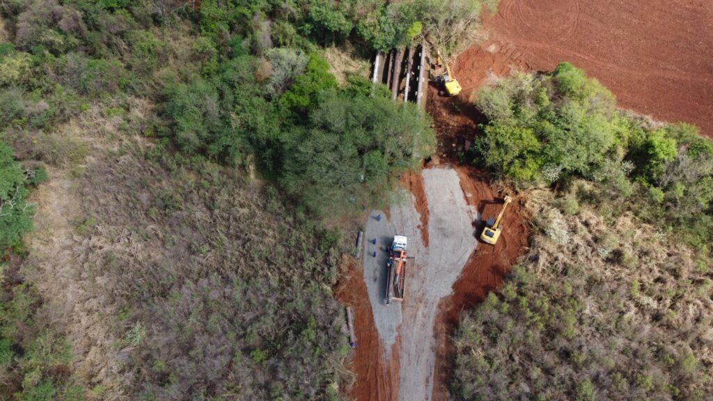 A imagem aérea mostra uma área de construção em andamento, com equipamentos pesados e trabalhadores. No centro da imagem, um caminhão branco com uma carreta laranja está parado em uma estrada de terra. Há várias pessoas ao redor do caminhão, algumas parecem estar descarregando materiais. À direita do caminhão, uma escavadeira amarela está em operação, com seu braço estendido em direção a uma área de terra vermelha. Mais adiante, outra escavadeira amarela está visível, também trabalhando em uma área de terra. Acima do caminhão, em direção ao topo da imagem, há uma estrutura de concreto com vários tubos de grande diâmetro passando por ela. Esta estrutura parece ser uma ponte ou um viaduto. A paisagem ao redor é uma mistura de vegetação densa, com árvores verdes e áreas de grama seca e arbustos. Há também uma grande área de terra arada, de cor avermelhada, em contraste com a vegetação. A presença de escavadeiras, um caminhão e trabalhadores indica que algum tipo de obra ou projeto de infraestrutura está sendo realizado no local. A estrutura com os tubos sugere que pode ser um projeto relacionado a água, esgoto ou transporte.