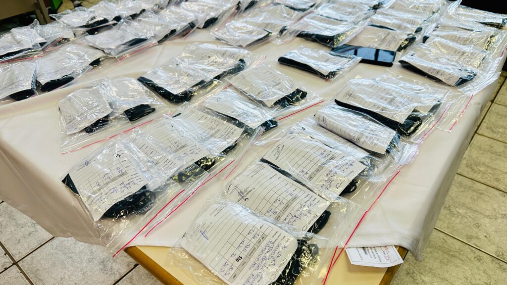 A imagem mostra uma mesa coberta com uma toalha branca, sobre a qual estão dispostos numerosos pacotes plásticos transparentes. Cada pacote contém um item escuro, possivelmente vestuário como meias, e uma folha de papel com anotações manuscritas. As anotações incluem o que parecem ser nomes, datas, números e outros detalhes, sugerindo um sistema de catalogação ou inventário. A disposição organizada dos pacotes em filas sugere que este é um processo de organização ou distribuição. O fundo da imagem mostra um piso com padrão de ladrilhos e uma parede.