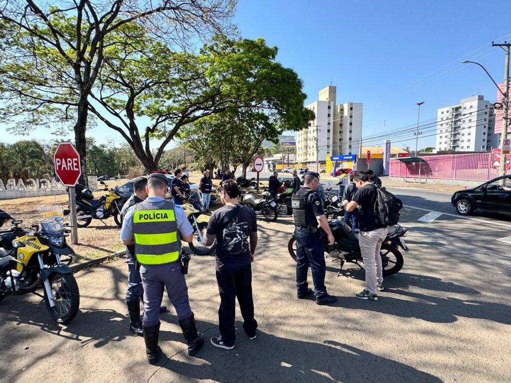 A imagem mostra um grupo de policiais em uniforme, alguns em motocicletas, reunidos em uma rua sob um céu azul e ensolarado. Há uma placa de "PARE" vermelha em primeiro plano, e árvores e edifícios ao fundo. Um policial, com um colete refletivo amarelo com a palavra "POLÍCIA" escrita em preto, está em primeiro plano, de costas para a câmera. Outros policiais estão em pé ou sentados em motocicletas, conversando entre si. As motocicletas são de cor amarela e preta, e algumas têm o logotipo "SMUTTRAN". A cena sugere uma operação policial ou uma parada de trânsito.