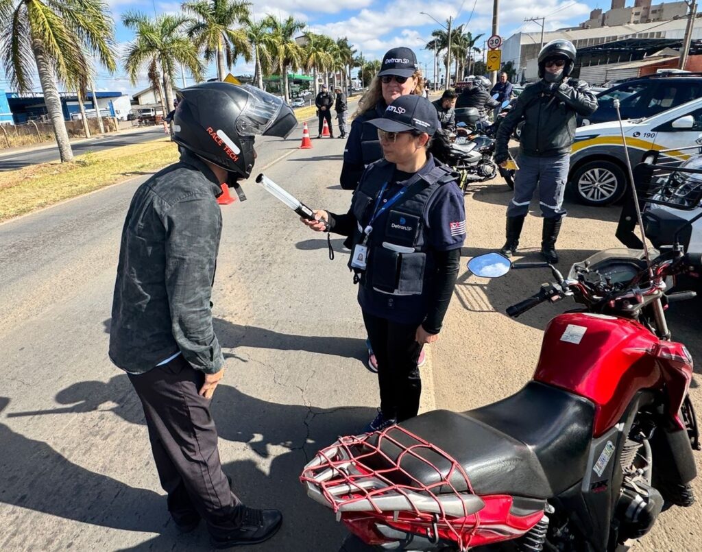 A imagem mostra uma operação de fiscalização de trânsito em uma via pública, possivelmente durante o dia, com céu azul e algumas nuvens. No primeiro plano, um homem com capacete preto, que tem a inscrição "RACE TECH" em vermelho, está em pé, de perfil, olhando para uma policial. Ele veste uma camisa de manga longa com estampa camuflada em tons de cinza e calças pretas. A policial, que usa um colete azul com o logo "Detran.SP" e um boné com a mesma inscrição, está em frente ao homem, segurando um bafômetro branco e apontando para ele, indicando que ele deve soprar. Ela também usa calças pretas. Ao fundo, outras pessoas vestidas com coletes do Detran.SP e bonés estão presentes, assim como motocicletas e carros de polícia. Uma mulher com óculos escuros e boné do Detran.SP está atrás da policial que está realizando o teste. Há também cones de sinalização laranja na pista.