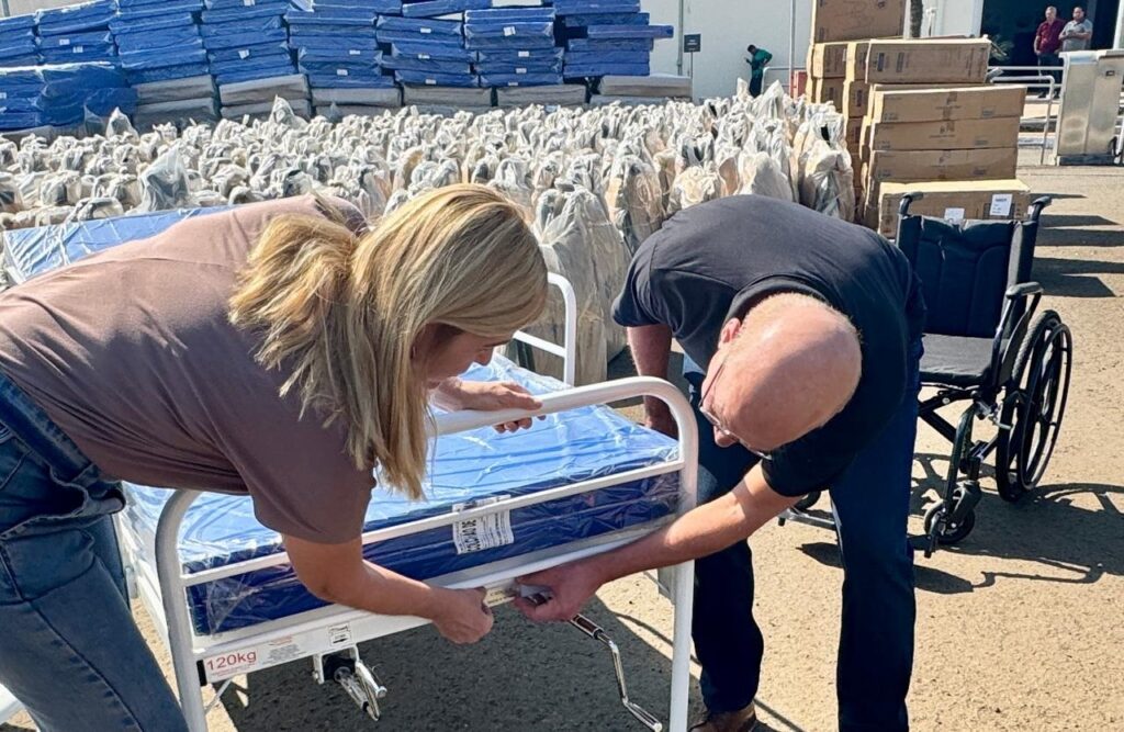 A imagem mostra duas pessoas, um homem e uma mulher, inclinados sobre uma cama de hospital. A mulher, com cabelos loiros e vestindo uma blusa marrom e jeans, está segurando a lateral da cama. O homem, careca e usando uma camiseta preta, está mexendo em um mecanismo na parte inferior da cama. Ao fundo, há uma grande quantidade de camas de hospital e colchões embalados, empilhados uns sobre os outros. Alguns dos colchões estão cobertos com plástico azul, enquanto outros estão embalados em material mais claro. Há também uma cadeira de rodas visível à direita da imagem. O cenário parece ser ao ar livre, com luz solar forte e sombras projetadas no chão. Em segundo plano, mais ao fundo, algumas caixas empilhadas e pessoas em pé podem ser vistas perto de um prédio.