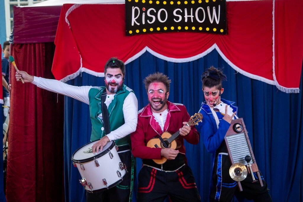 A imagem mostra três palhaços em um palco com uma cortina vermelha e azul ao fundo. Acima deles, há uma placa amarela com a inscrição "RISO SHOW" em letras pretas. O palhaço da esquerda está vestindo um colete verde sobre uma camisa branca e toca um tambor branco com duas baquetas. Ele tem maquiagem de palhaço com um nariz vermelho e detalhes brancos no rosto. O palhaço do meio está usando uma jaqueta vermelha com detalhes prateados e uma camisa branca com babados. Ele segura um pequeno violão e tem uma expressão facial animada, com a boca aberta em um grito. Sua maquiagem é semelhante à do outro palhaço. O palhaço da direita veste um casaco azul com detalhes brancos e pretos. Ele está tocando uma tábua de lavar com um pequeno prato e um apito, e faz um gesto com a mão. Sua maquiagem também inclui um nariz vermelho e detalhes brancos. Todos os palhaços parecem estar se apresentando em um show.