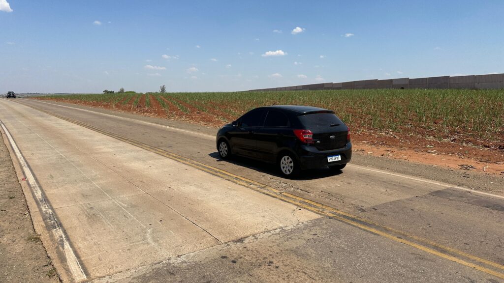 A imagem mostra uma estrada de asfalto sob um céu azul claro com algumas nuvens brancas. À direita da estrada, há um campo de cultivo com fileiras organizadas de plantas verdes e solo vermelho. No campo, ao fundo, há uma longa fileira de estruturas retangulares de concreto, possivelmente um muro ou uma barragem. Na estrada, um carro preto de quatro portas está em movimento, andando na mesma direção que a câmera. O carro está na faixa da direita, e a câmera parece estar em um veículo que está ultrapassando o carro preto pela esquerda. A estrada tem marcações de faixa amarelas duplas e uma faixa branca na borda esquerda. A paisagem ao redor sugere um ambiente rural ou agrícola, possivelmente em um dia ensolarado e quente. A cultura no campo parece ser de cana-de-açúcar ou algo semelhante, com os brotos verdes emergindo do solo avermelhado. A fileira de estruturas de concreto adiciona um elemento incomum à paisagem agrícola.