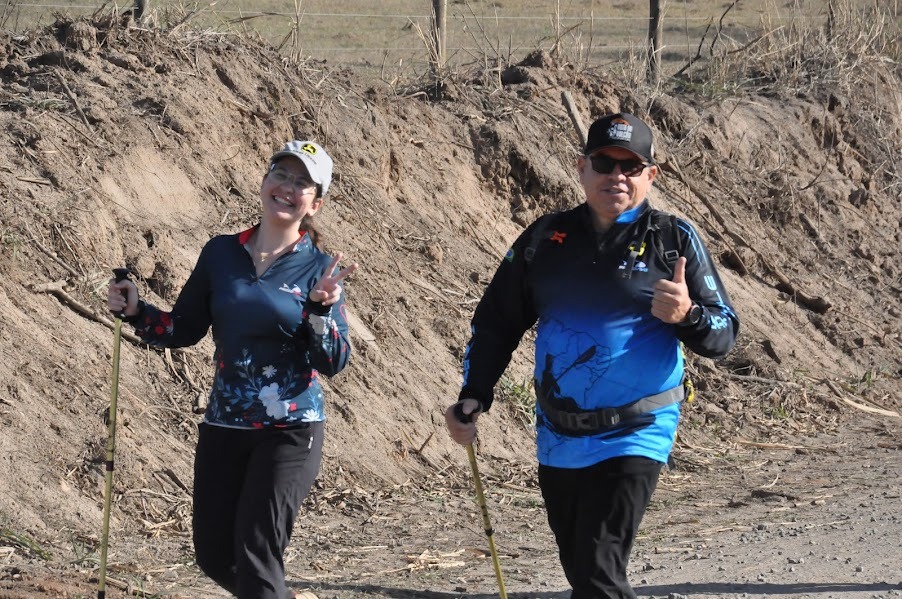 A mulher, à esquerda, usa um boné branco com o logo da John Deere, óculos e uma blusa de manga comprida azul com estampa floral. Ela segura um bastão de caminhada em sua mão esquerda e faz o sinal de paz com a mão direita.O homem, à direita, usa um boné preto, óculos escuros e uma blusa de manga comprida azul com um degradê e uma estampa de caiaque. Ele também segura um bastão de caminhada em sua mão direita e exibe um joinha com a mão esquerda.Ambos estão vestidos com roupas esportivas adequadas para a atividade e parecem estar em um caminho de terra com uma encosta de terra e vegetação seca ao fundo. A atmosfera geral da imagem é de alegria e atividade ao ar livre.