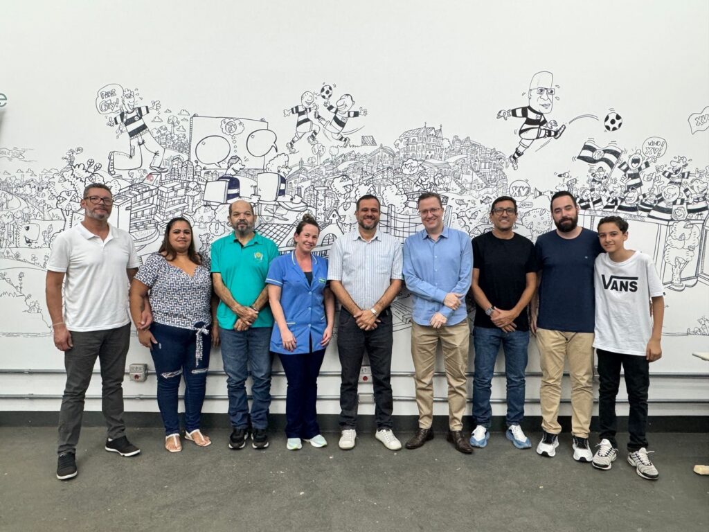 A imagem mostra um grupo de dez pessoas, incluindo homens, mulheres e um jovem, posando em frente a uma parede com um mural elaborado em preto e branco. O mural retrata uma cena urbana com vários elementos, incluindo prédios, pessoas em movimento e um jogo de futebol, com personagens caricaturados em destaque. As pessoas estão alinhadas e olhando para a câmera. A iluminação é clara, destacando os indivíduos e o mural. A composição sugere um evento ou encontro em um ambiente interno, possivelmente um escritório ou espaço comunitário, decorado com arte.