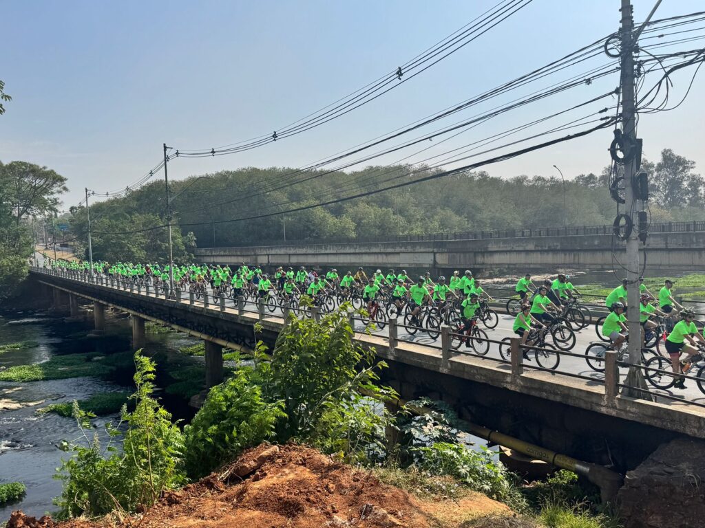 A imagem mostra um grande grupo de ciclistas percorrendo uma ponte em um dia ensolarado.Os ciclistas vestem camisetas verdes vibrantes e capacetes, sugerindo que fazem parte de um evento organizado ou passeio em grupo. A ponte sobre a qual eles pedalam parece ser de concreto e é sustentada por pilares. Abaixo da ponte, um rio ou córrego flui, com vegetação crescendo em suas margens.Ao fundo, há uma linha de árvores exuberantes e uma estrada elevada, com postes de eletricidade e fios atravessando o céu. O céu está claro, indicando um dia limpo e provavelmente quente. A perspectiva da foto é de baixo para cima, dando uma sensação de grandiosidade ao evento e à paisagem. A composição geral transmite uma sensação de atividade comunitária, saúde e apreciação da natureza.