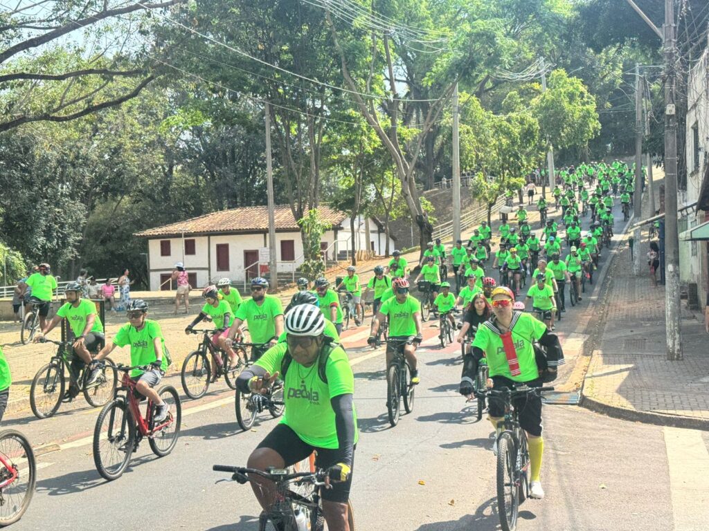 A imagem mostra um grande grupo de ciclistas, a maioria vestindo camisetas verde-limão com o logotipo "Pedala". Eles estão pedalando em uma rua ensolarada, com árvores e edifícios ao redor.Os ciclistas estão em uma formação em leque, com alguns na frente e outros mais atrás. A maioria parece estar participando de um evento ou passeio ciclístico. A rua é inclinada, com um leve aclive, e há pessoas observando o grupo em ambos os lados da rua.A vegetação exuberante e as árvores frondosas sugerem que o evento está ocorrendo em uma área com bastante natureza. O céu está claro e azul, indicando um dia ensolarado.Em primeiro plano, há vários ciclistas mais próximos à câmera, permitindo ver detalhes de suas roupas e bicicletas. Um ciclista em particular, na extrema direita, usa óculos de sol coloridos e uma bandana.No geral, a imagem transmite uma sensação de comunidade e atividade física, com um grupo de pessoas desfrutando de um passeio de bicicleta em um ambiente agradável.
