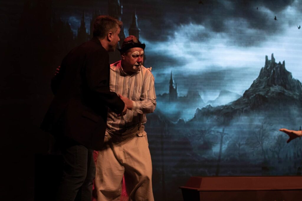 A imagem mostra uma cena teatral com dois homens e um cenário sombrio ao fundo. À esquerda, um homem vestido com um terno escuro parece estar conversando com o homem à sua direita. Este último está vestido como um palhaço, com maquiagem branca e vermelha no rosto, um chapéu e uma camisa listrada com calças largas e claras. O fundo é uma projeção digital de uma paisagem sombria e fantasmagórica, com montanhas escarpadas, castelos em ruínas e névoa, evocando uma atmosfera gótica ou de terror. No primeiro plano, à direita, parte de um caixão marrom é visível. Uma mão estendida, possivelmente de um terceiro ator, aponta para a paisagem. A iluminação sugere um palco, com foco nos atores e no cenário.