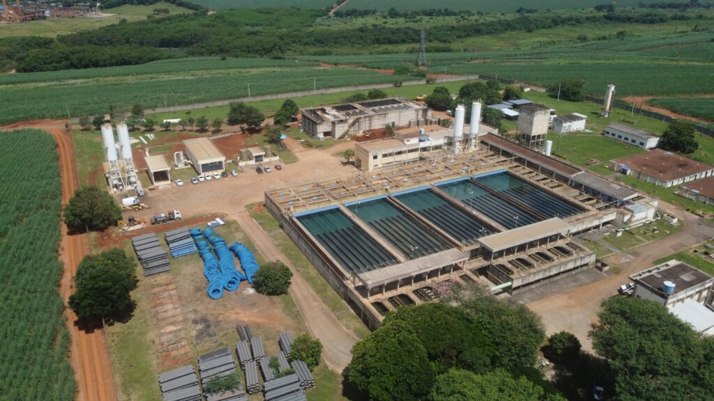 A imagem aérea mostra uma grande estação de tratamento de água, com vários tanques retangulares de água verde, dispostos em fileiras. Ao redor da estação, há vários edifícios industriais, silos e torres. À esquerda da estação de tratamento, há um grande campo de cana-de-açúcar, com uma estrada de terra que leva a ela. Há também pilhas de tubos azuis e cinzas, possivelmente para uso na estação ou em projetos relacionados. Ao fundo, a paisagem é composta por mais campos verdes e algumas áreas de floresta, com uma torre de transmissão de energia visível à distância. O céu está claro e o dia parece ensolarado.