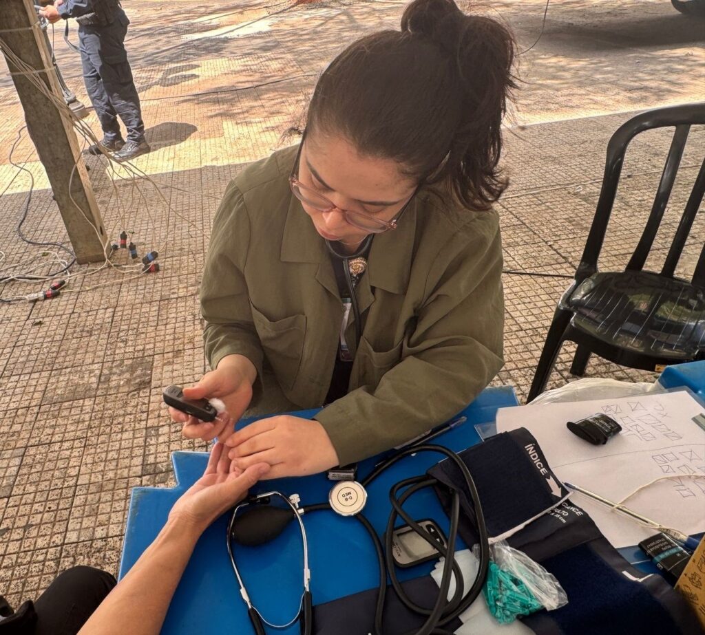 A imagem mostra uma profissional de saúde, possivelmente uma enfermeira ou técnica de enfermagem, realizando um teste de glicemia capilar em uma pessoa. A profissional, usando óculos e um jaleco verde, segura um glicosímetro e um lancetador, pronto para coletar uma amostra de sangue do dedo da paciente. Na mesa azul à frente, há diversos materiais médicos, incluindo um estetoscópio, um esfigmomanômetro (aparelho de pressão arterial), tiras de teste de glicemia e um pequeno recipiente com lancetas. Ao fundo, em uma área externa com piso de ladrilhos, observam-se cabos elétricos e uma pessoa vestida com uniforme, possivelmente um policial ou segurança, em pé. Uma cadeira preta de plástico está visível no canto direito da imagem. A cena sugere um ambiente de atendimento médico comunitário ou de triagem.