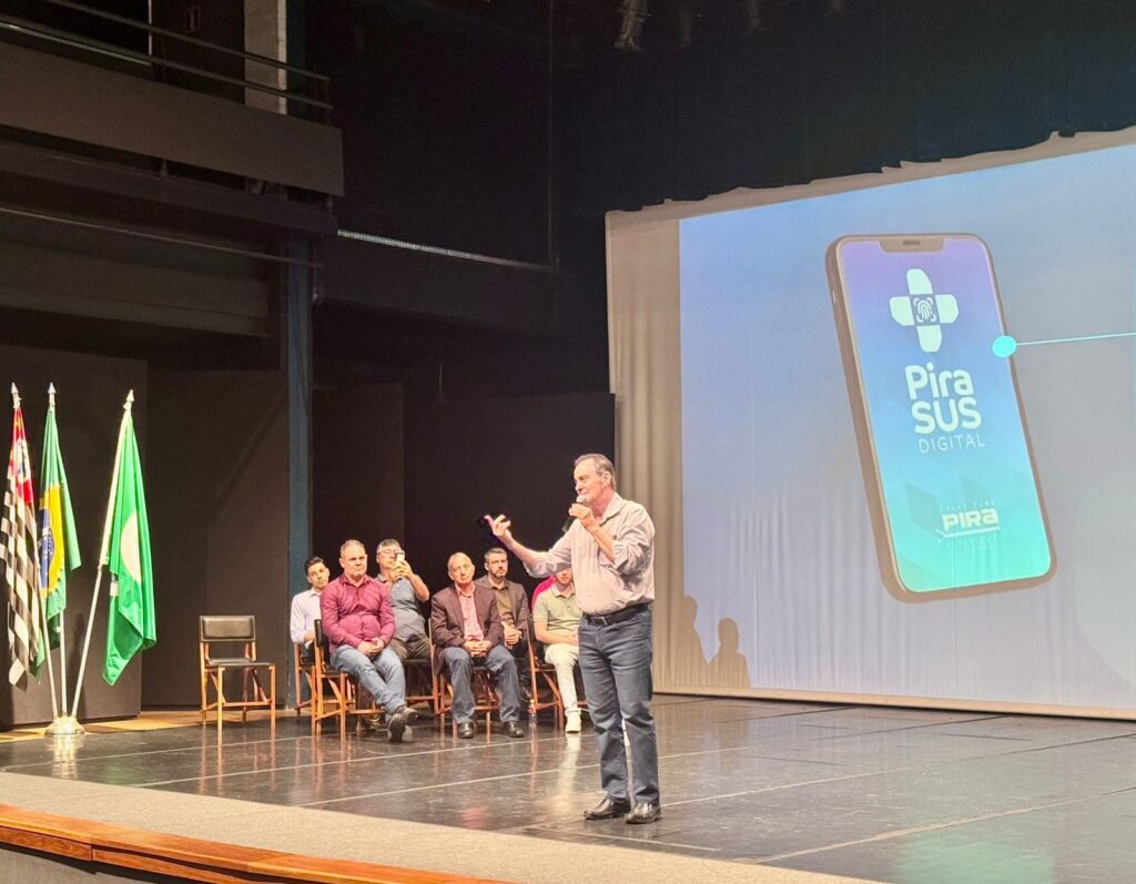 A imagem mostra o prefeito de Piracicaba, Helinho Zanatta, falando em um microfone em um palco, com um telão ao fundo exibindo um aplicativo de celular chamado "Pira SUS Digital". O homem está vestido com uma camisa de manga comprida e calças jeans, e gesticula com a mão enquanto fala. Ao fundo, há um grupo de homens sentados em cadeiras, e à esquerda, três bandeiras. O palco é escuro, com iluminação focada no homem e no telão.