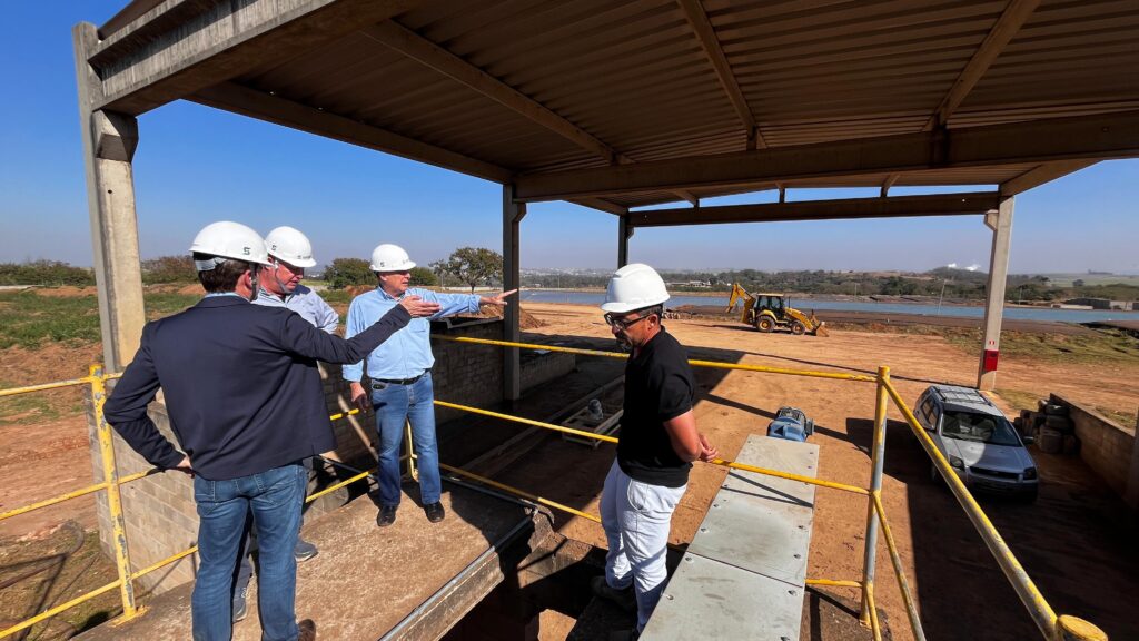 O grupo de homens está em um canteiro de obras, sob uma estrutura de metal com um telhado. Todos usam capacetes brancos. Um homem de terno azul escuro e calça jeans está de costas para a câmera, com a mão direita apontando para a frente. Ao seu lado, um homem com camisa azul clara e calça jeans também aponta com a mão direita. Outro homem, com camisa cinza e calça jeans, está em pé entre eles. Um quarto homem, de camiseta preta e calça branca, está em primeiro plano, virado para a direita. Ao fundo, vê-se uma paisagem com um lago, uma retroescavadeira amarela e alguns prédios ao longe. O céu está claro e azul. Há uma pequena estrutura de concreto com uma escada e um corrimão amarelo em primeiro plano. Um carro prateado está estacionado à direita.