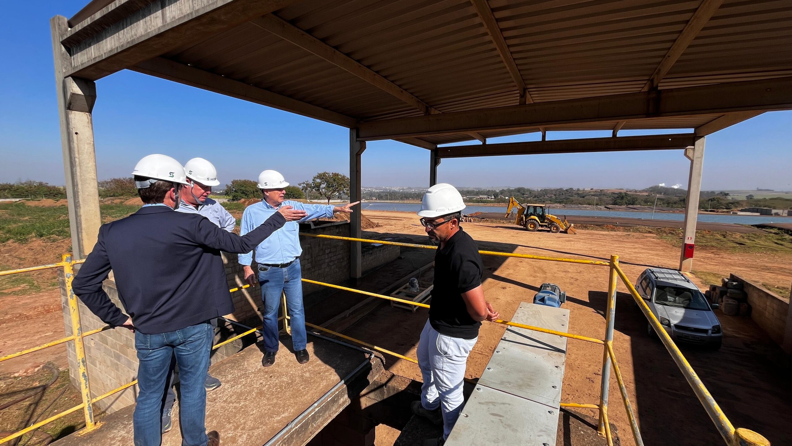 O grupo de homens está em um canteiro de obras, sob uma estrutura de metal com um telhado. Todos usam capacetes brancos. Um homem de terno azul escuro e calça jeans está de costas para a câmera, com a mão direita apontando para a frente. Ao seu lado, um homem com camisa azul clara e calça jeans também aponta com a mão direita. Outro homem, com camisa cinza e calça jeans, está em pé entre eles. Um quarto homem, de camiseta preta e calça branca, está em primeiro plano, virado para a direita. Ao fundo, vê-se uma paisagem com um lago, uma retroescavadeira amarela e alguns prédios ao longe. O céu está claro e azul. Há uma pequena estrutura de concreto com uma escada e um corrimão amarelo em primeiro plano. Um carro prateado está estacionado à direita.