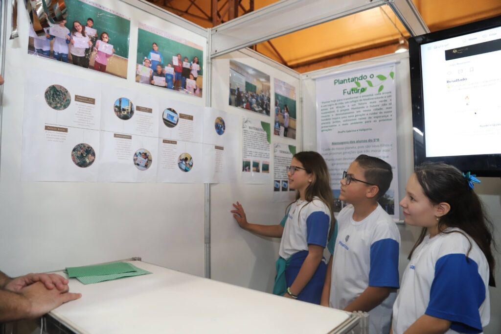 A imagem mostra um grupo de crianças em uma feira de ciências, apresentando um projeto. Três crianças estão em primeiro plano, olhando para um painel com informações sobre o projeto "Plantando o Futuro". O painel inclui fotos de alunos, mapas e textos explicativos. Ao fundo, outros painéis e fotos de atividades escolares. Há também um monitor exibindo uma plataforma de quiz. As crianças vestem uniformes escolares. O projeto, intitulado "Plantando o Futuro", parece focar em sustentabilidade e meio ambiente, com mensagens de alunos sobre a importância de cuidar do planeta para as futuras gerações. Um dos textos no painel cita os professores Sabrina e Valquiria. A feira de ciências parece ser um evento escolar onde os alunos apresentam seus trabalhos e aprendizados.