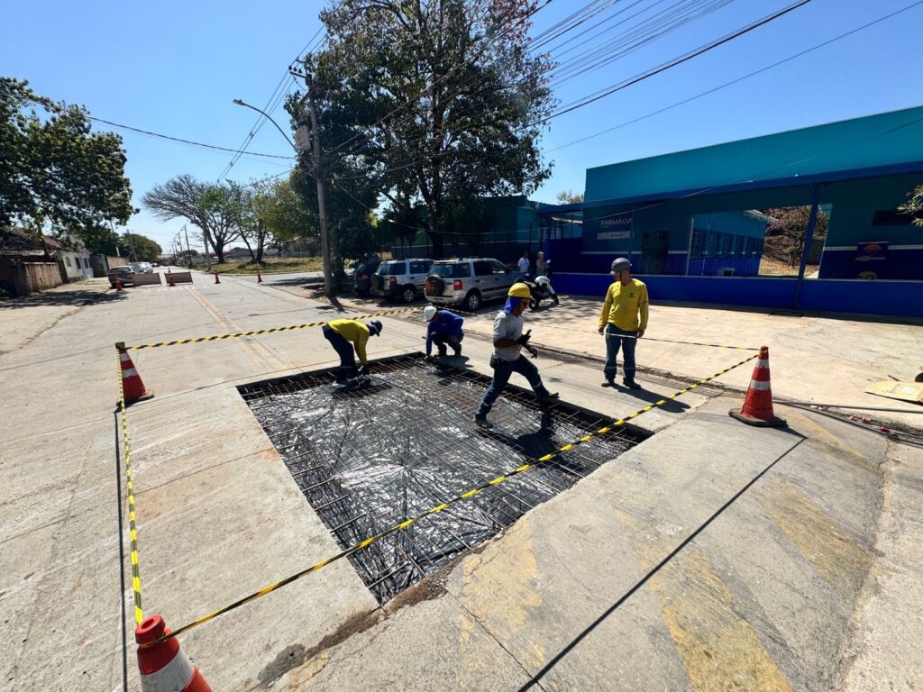 A imagem mostra uma equipe de trabalhadores em uma rua, realizando obras em uma grande vala aberta no asfalto. A vala está forrada com uma manta prateada e uma malha de vergalhões, indicando que uma nova estrutura de concreto está sendo preparada. Vários trabalhadores, todos usando capacetes de segurança, estão concentrados na tarefa. Um deles, vestindo uma camisa amarela, parece supervisionar o trabalho, enquanto outros dois estão agachados dentro da vala, possivelmente ajustando a armação de metal. Outro trabalhador, também de amarelo, está em pé na borda da vala. A área da obra está isolada com cones de trânsito laranja e fita zebrada amarela e preta, para alertar os motoristas e pedestres sobre o perigo. Ao fundo, carros estão estacionados na rua, e um prédio azul com a palavra "FARMÁCIA" escrita em branco é visível. O céu está claro e azul, sugerindo um dia ensolarado.