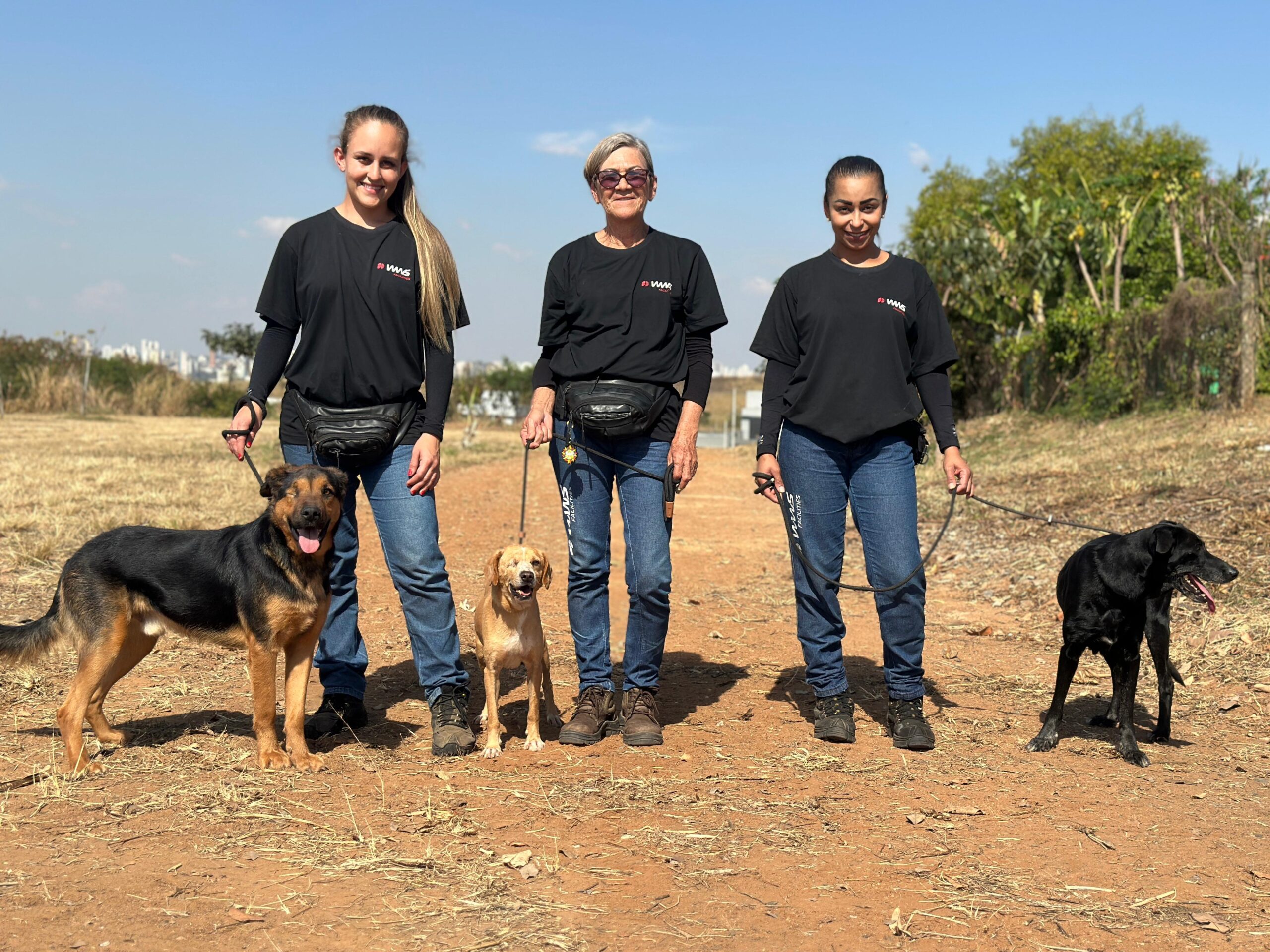 Na imagem, três mulheres estão em pé em um campo aberto, cada uma com um cachorro na coleira. Todas as mulheres vestem camisetas pretas com um logotipo vermelho e branco no lado esquerdo do peito, calças jeans azuis e pochetes pretas na cintura. A mulher da esquerda, com cabelos loiros longos presos em um rabo de cavalo, está segurando a coleira de um cachorro grande, de pelo preto e marrom. O cachorro está olhando para a câmera com a língua para fora. A mulher do meio, mais velha, usa óculos escuros e tem cabelos curtos e grisalhos. Ela está segurando a coleira de um cachorro de porte médio, de cor caramelo. O cachorro também está olhando para a câmera, com a boca aberta e a língua para fora. A mulher da direita tem cabelos escuros presos para trás. Ela está segurando a coleira de um cachorro preto, que parece ser um Labrador. O cachorro está olhando para a direita, com a língua para fora. O fundo da imagem mostra um campo seco com algumas árvores e arbustos ao longe, sob um céu azul claro. Uma cidade com edifícios pode ser vista ao fundo, à esquerda. O chão em primeiro plano é de terra batida com algumas folhas secas.