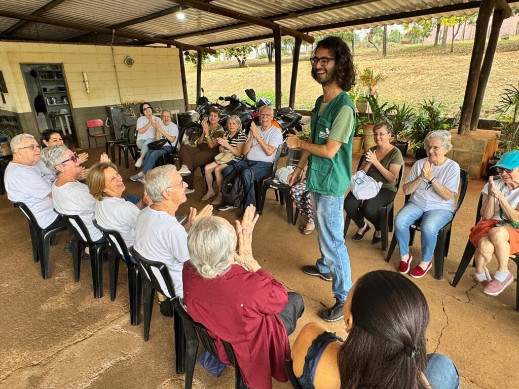 A imagem mostra um grupo de idosos sentados em cadeiras, com um homem em pé à frente deles. O homem, vestindo um colete verde, parece estar falando ou apresentando algo para o grupo. A maioria dos idosos está sorrindo e aplaudindo, indicando um ambiente positivo e engajado. O cenário parece ser ao ar livre, sob uma estrutura coberta, com vegetação e árvores ao fundo. A iluminação sugere que é dia.