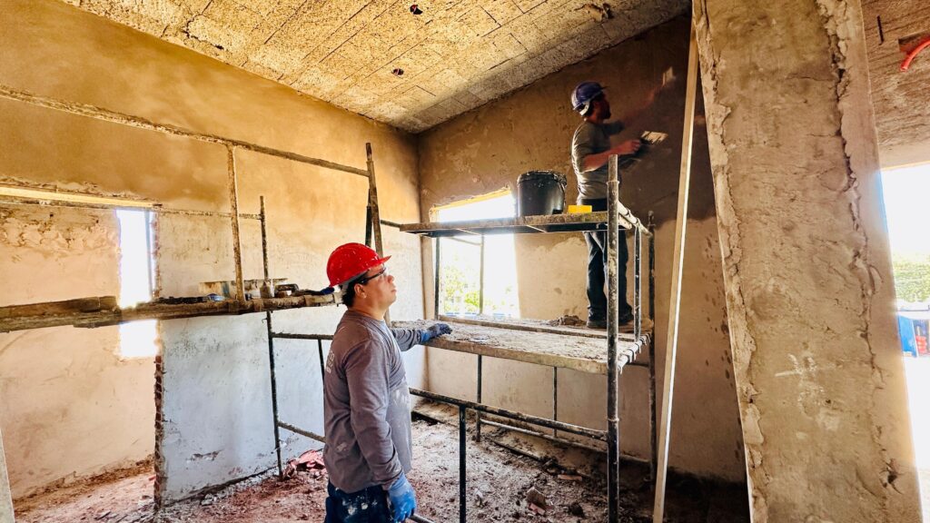 A imagem mostra dois trabalhadores realizando o reboco de um cômodo em construção. No primeiro plano, à esquerda, um trabalhador com capacete vermelho e óculos olha para cima, para o teto, enquanto segura uma ferramenta. Ele está em pé no chão do cômodo, que está coberto de entulho e materiais de construção. Ao fundo, à direita, outro trabalhador está em cima de um andaime, aplicando reboco em uma parede. Ele usa um capacete azul e está concentrado em seu trabalho. O andaime está posicionado de forma a dar acesso à parte superior das paredes e ao teto. O cômodo em si parece estar em um estágio inicial de acabamento. As paredes estão com o reboco fresco, com texturas irregulares em algumas áreas. O teto é aparente, com um material que parece ser concreto ou gesso. Há aberturas nas paredes que parecem ser janelas ou vãos, permitindo a entrada de luz natural. O chão está sujo, com pedaços de reboco, poeira e outros detritos de construção. O ambiente geral sugere um local de trabalho ativo e em progresso.