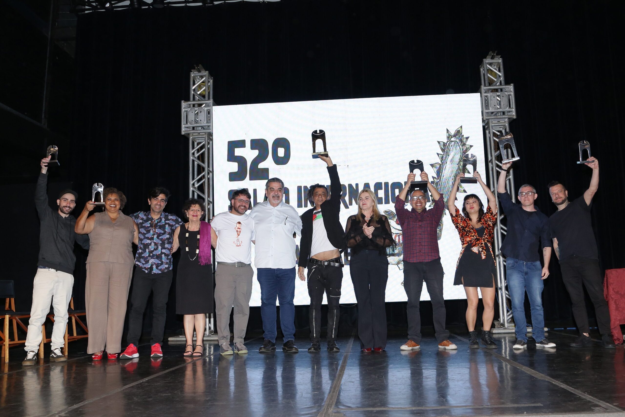Grupo de artistas premiados posa no palco segurando troféus do Salão de Humor.