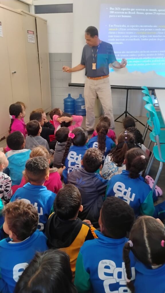 A imagem mostra uma sala de aula onde um homem está em pé em frente a um projetor, apresentando algo para um grupo de crianças sentadas no chão. O homem, vestindo uma camisa azul e calças bege, gesticula com as mãos enquanto fala. O projetor exibe texto em uma tela branca, com alguns pontos destacados. As crianças, vestidas com uniformes escolares azuis com o logo "Edu Ca", estão sentadas em círculo, olhando atentamente para o apresentador. Algumas crianças estão com as mãos levantadas ou apoiadas no chão. Ao fundo, há armários e garrafões de água. A cena sugere uma atividade educativa ou uma palestra para crianças.